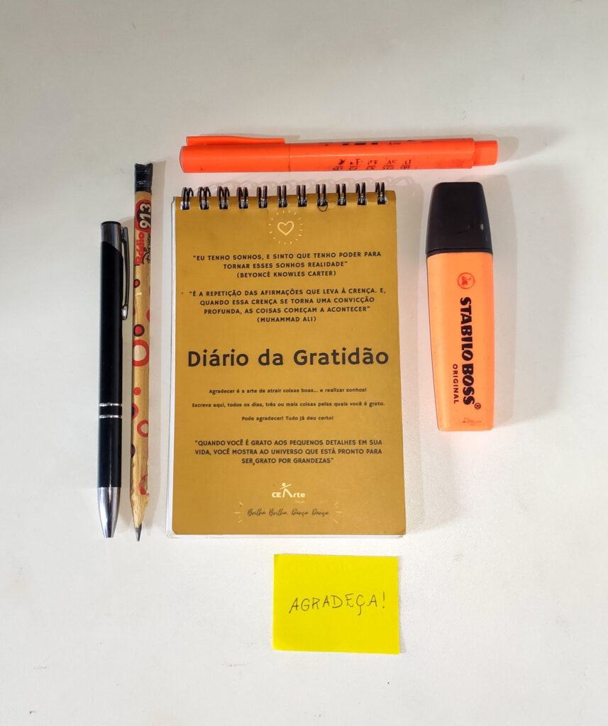 Foto com um bloco de notas bege, escrito em sua capa "Diário da Gratidão". O fundo da imagem é branco, e em volta do bloco contém alguns elementos. Acima e ao lado direito, marce-textos laranjas. Ao lado esquerdo, uma caneta preta e um lápis bege. Abaixo, um post-it amarelo, escrito "AGRADEÇA!".