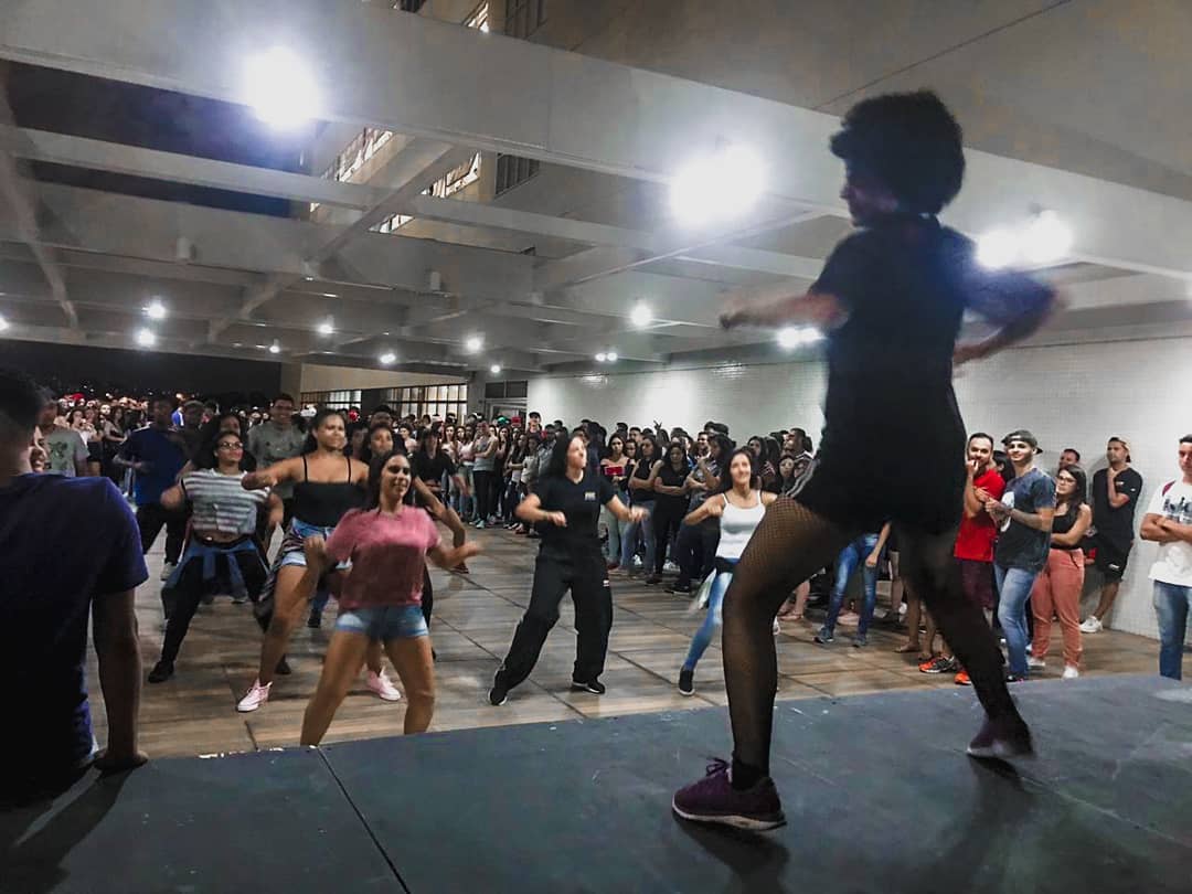 Uma mulher em cima de um palco dando aula de dança para várias pessoas.