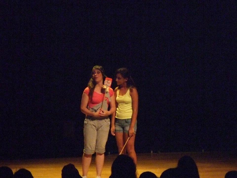 Duas meninas em cena, em cima de um palco, conversando.