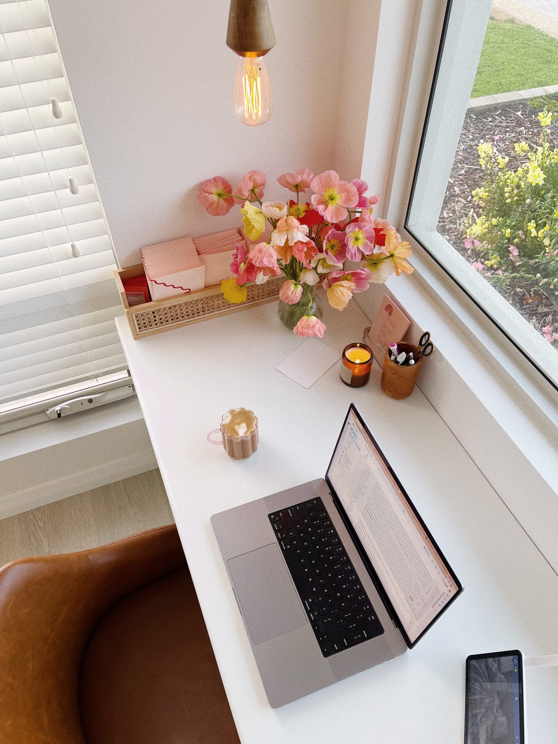 Mesa branca vista de cima. Em cima dela estão um computador (notebook) aberto e um celular. Ao canto, um arranjo de flores, itens de papelaria e uma vela, tudo nas cores rosa e amarelo.