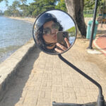 selfie minha, tirada no espelho de uma moto elétrica, na beira da praia.