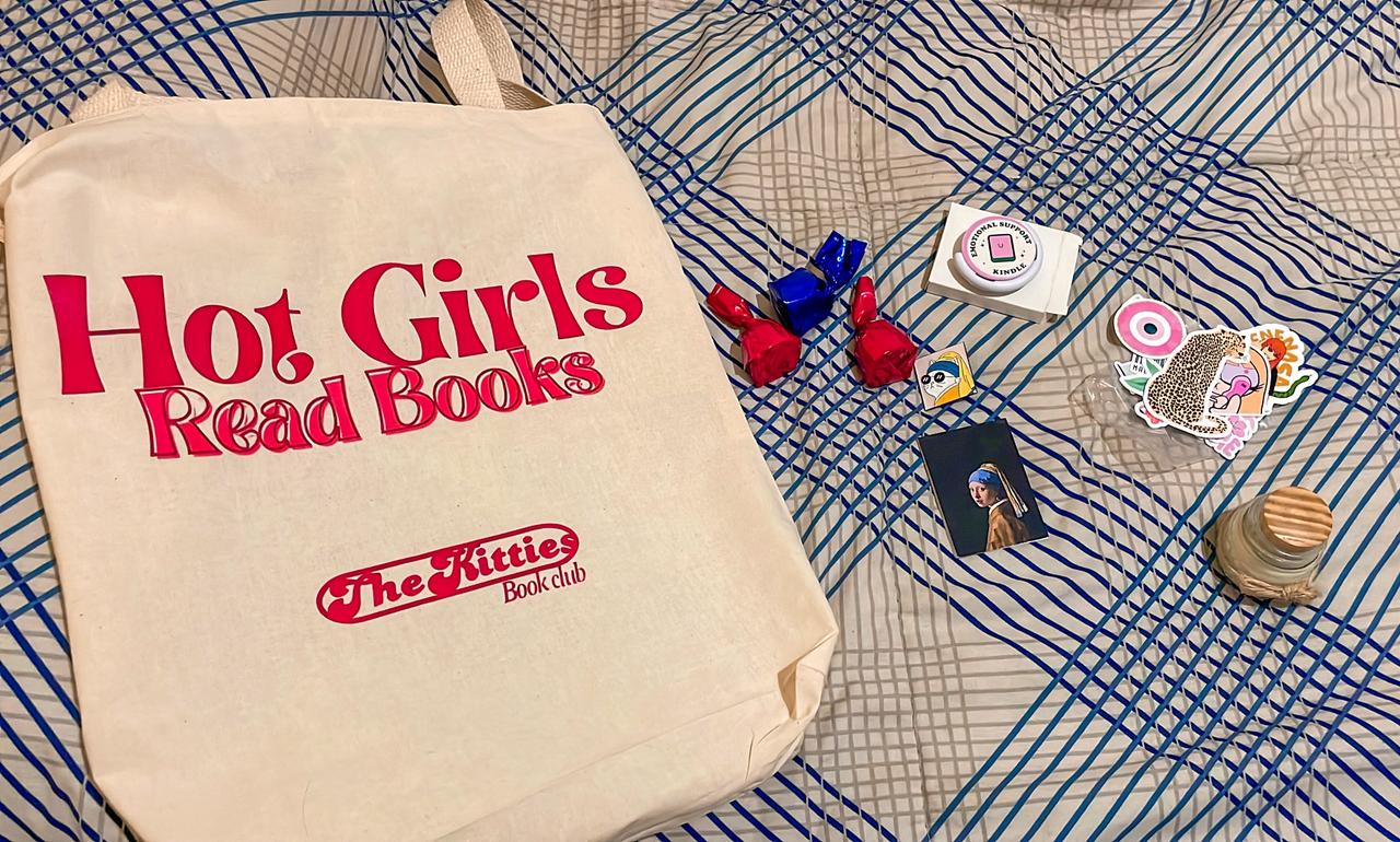 foto dos mimos do clube do livro: uma ecobag branca escrita "hot girls read books - the kitties book club", bombons, um pop socket para kindle, um pin, um marca-páginas, alguns adesivos e uma vela.
