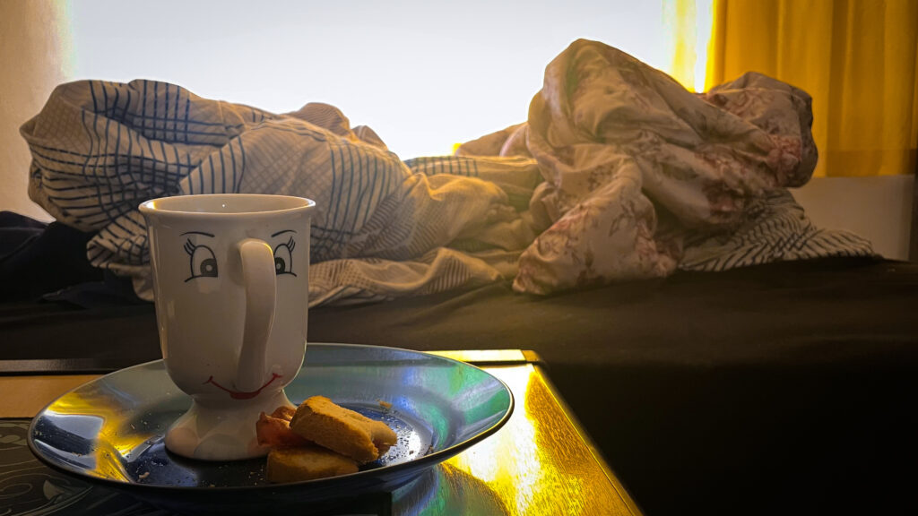 foto da minha caneca de café do Zip (personagem de A Bela e a Fera), com um último pedaço de pão, e a cama desarrumada ao fundo