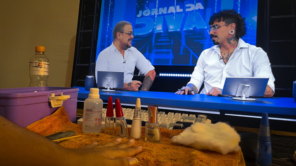 foto da mesa com esmaltes em cima e uma das minhas mãos apoiada, pronta para iniciar o processo de esmaltação. ao fundo, a tela do computador com o Fih e o Edu, do canal Diva Depressão.