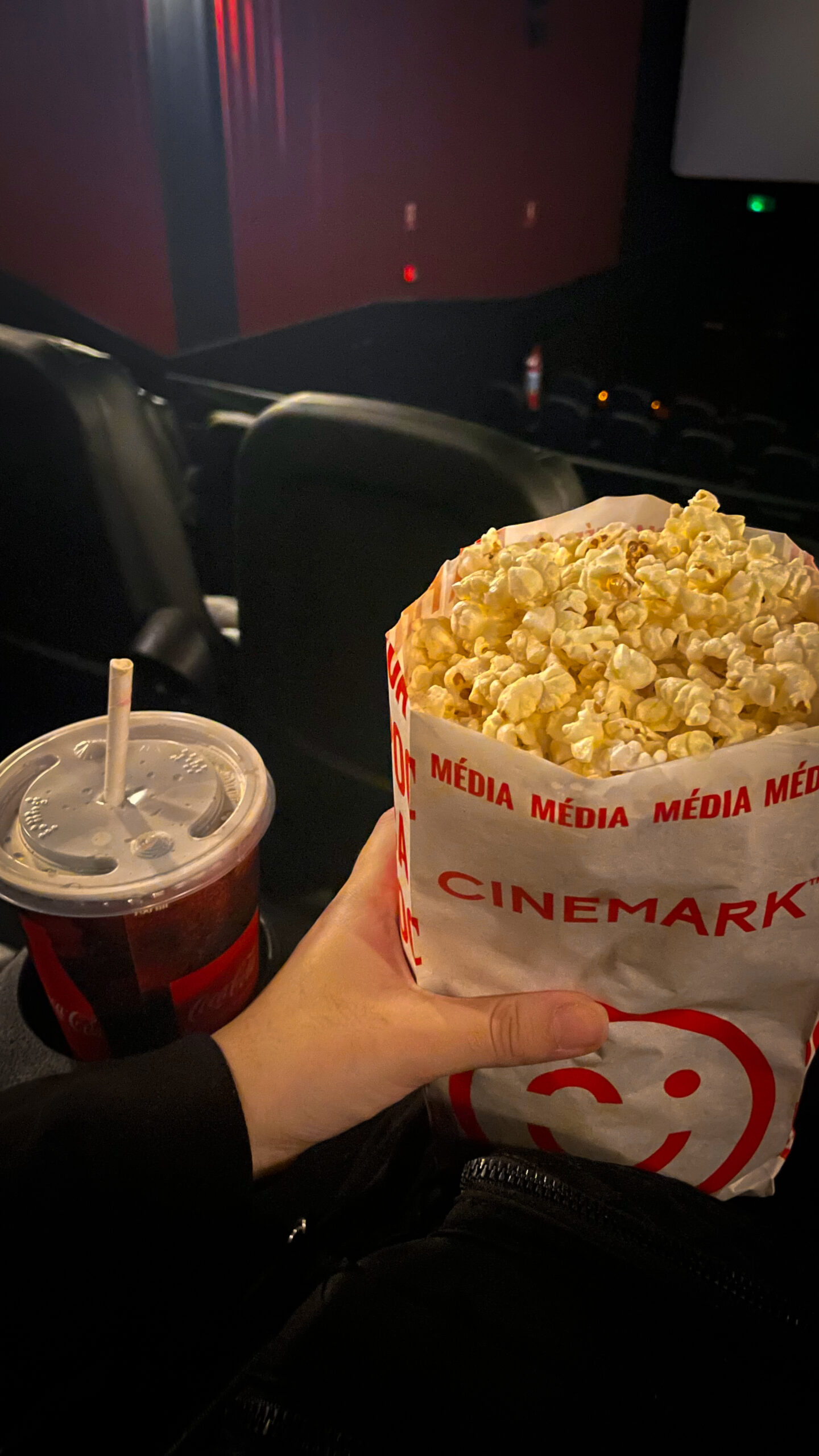 foto no cinema, vista do copo de refrigerante apoiado no braço da poltrona e uma mão segurando um saco de pipoca da Cinemark