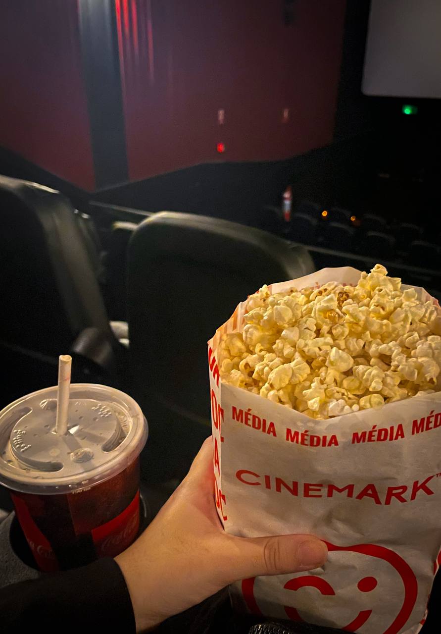 foto do cinema. vista da minha mão segurando um saco de pipoca e um copo de refigerante ao lado, apoiado no suporte da cadeira.