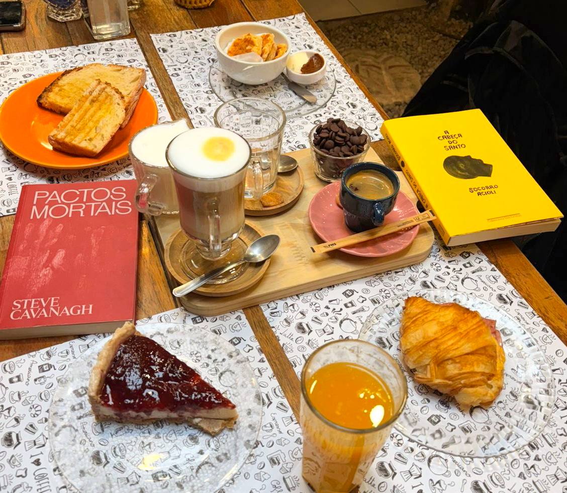 Mesa de café, com diversas xícaras e cafés, e dois livros em cima: A Cabeça do Santo do lado direito da mesa, e Pactos Mortais do lado esquerdo
