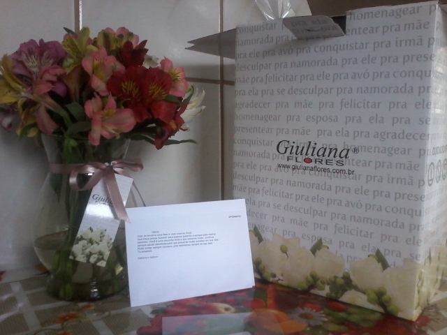 Foto de um jarro com flores, uma caixa da Giuliana Flores ao lado e uma carta.