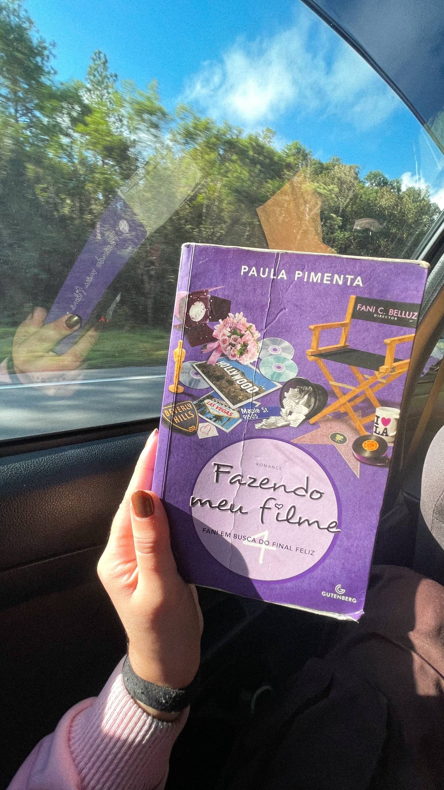 Mão segurando o livro "Fazendo Meu Filme 4" em frente a janela de um carro em movimento, que mostra árvores do lado de fora.