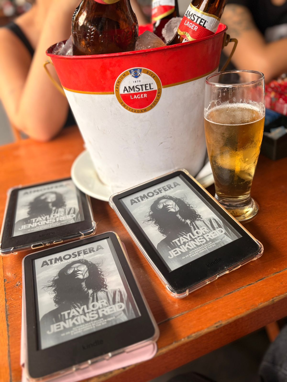 mesa do bar onde foi o encontro, com 3 kindles mostrando a capa do livro "Atmosfera", um copo de cerveja ao lado e um balde com garrafas de cerveja ao fundo
