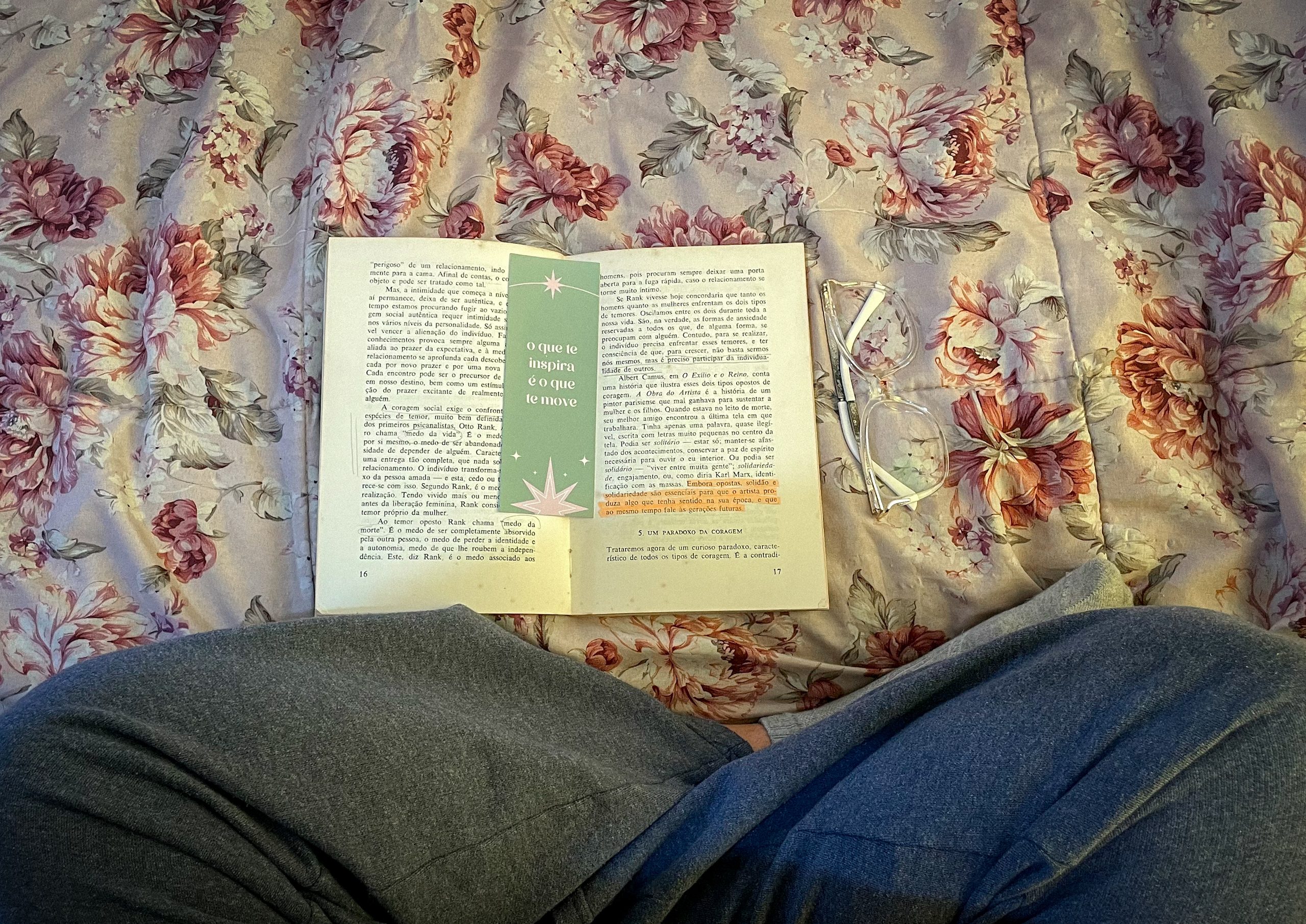 Livro aberto em cima da cama, com um óculos ao lado e minhas pernas cruzadas na frente. Dentro do livro, um marca páginas escrito "o que te inspira é o que te move"