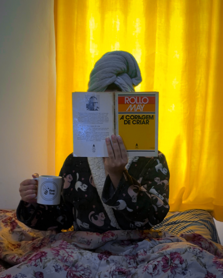 eu, sentada na cama, com uma coberta cobrindo minhas pernas, que estão cruzadas. uso roupão e estou segurando uma xícara numa mão e o livro na outra. o livro está na altura do meu rosto, cobrindo-o. utilizo uma toalha enrolada na cabeça e, ao fundo, a cortina amarela traz iluminação à foto.