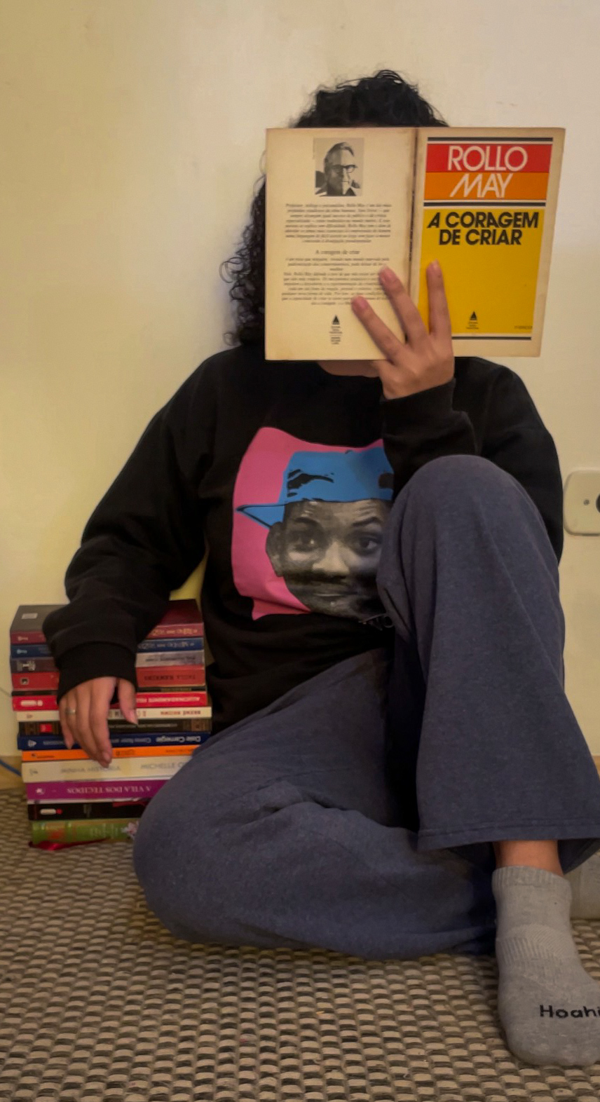 eu, sentada no chão, lendo o livro (que cobre meu rosto), com o outro braço apoiado em uma pilha de livros.