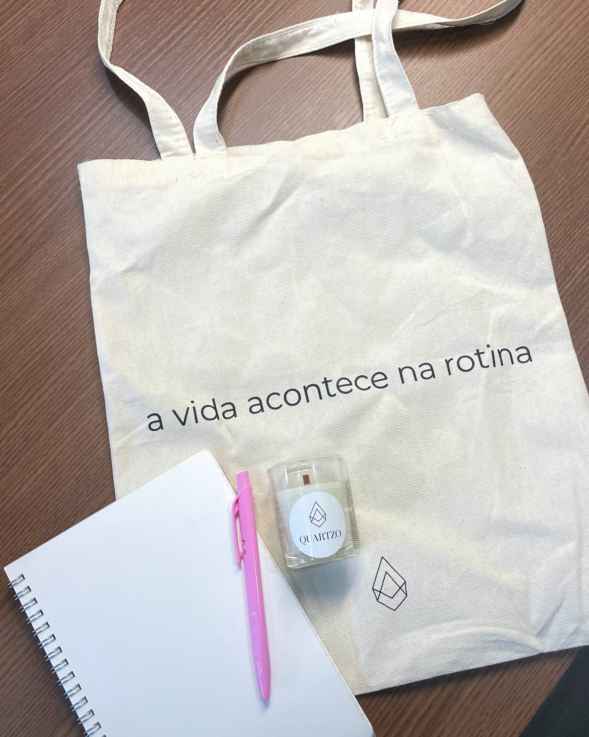 itens em cima de uma mesa vistos de cima: ecobag branca com o texto "a vida acontece na rotina", vela artesanal, caneta cor de rosa e um caderninho de capa branca