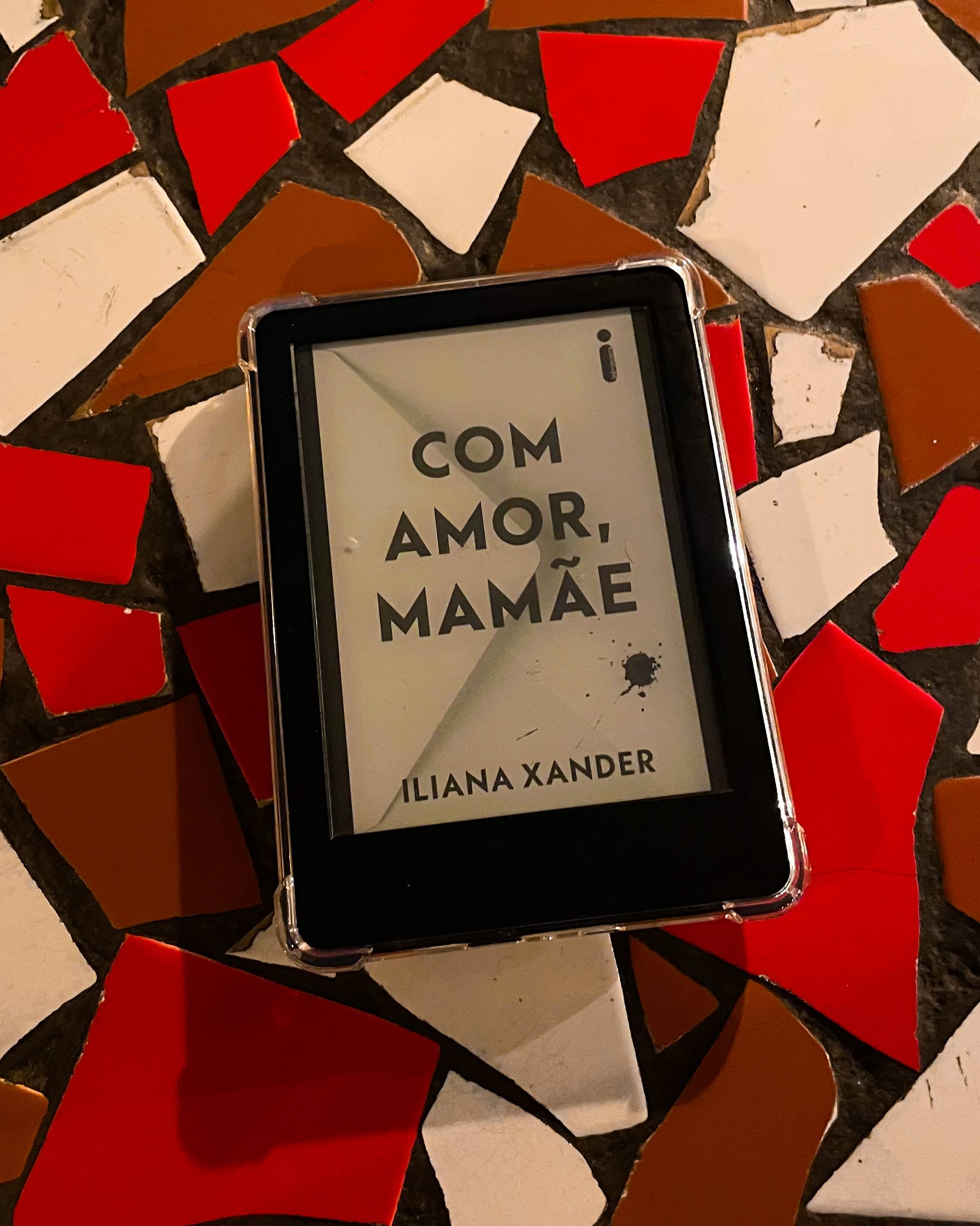 Foto de um kindle exibindo o livro "Com amor, mamãe" na capa. Apoiado em uma mesa revestida com caquinhos vermelhos, marrons e brancos.
